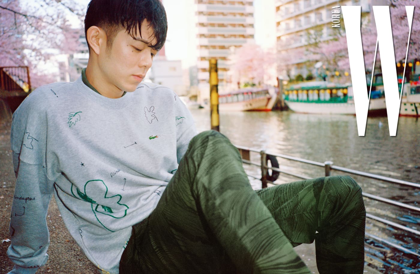 그레이 색상의 트로피컬 프린트 스웨트셔츠와 안에 겹쳐 입은 카키색 피케 폴로 셔츠, 나무 질감을 표현한 프린트가 독특한 치노 팬츠는 Lacoste Live 제품.