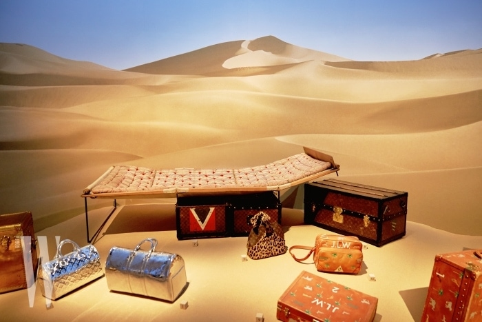 사막 한가운데에 놓인 듯한 연출이 인상적인 트렁크와 여행 가방, 간이 침대.